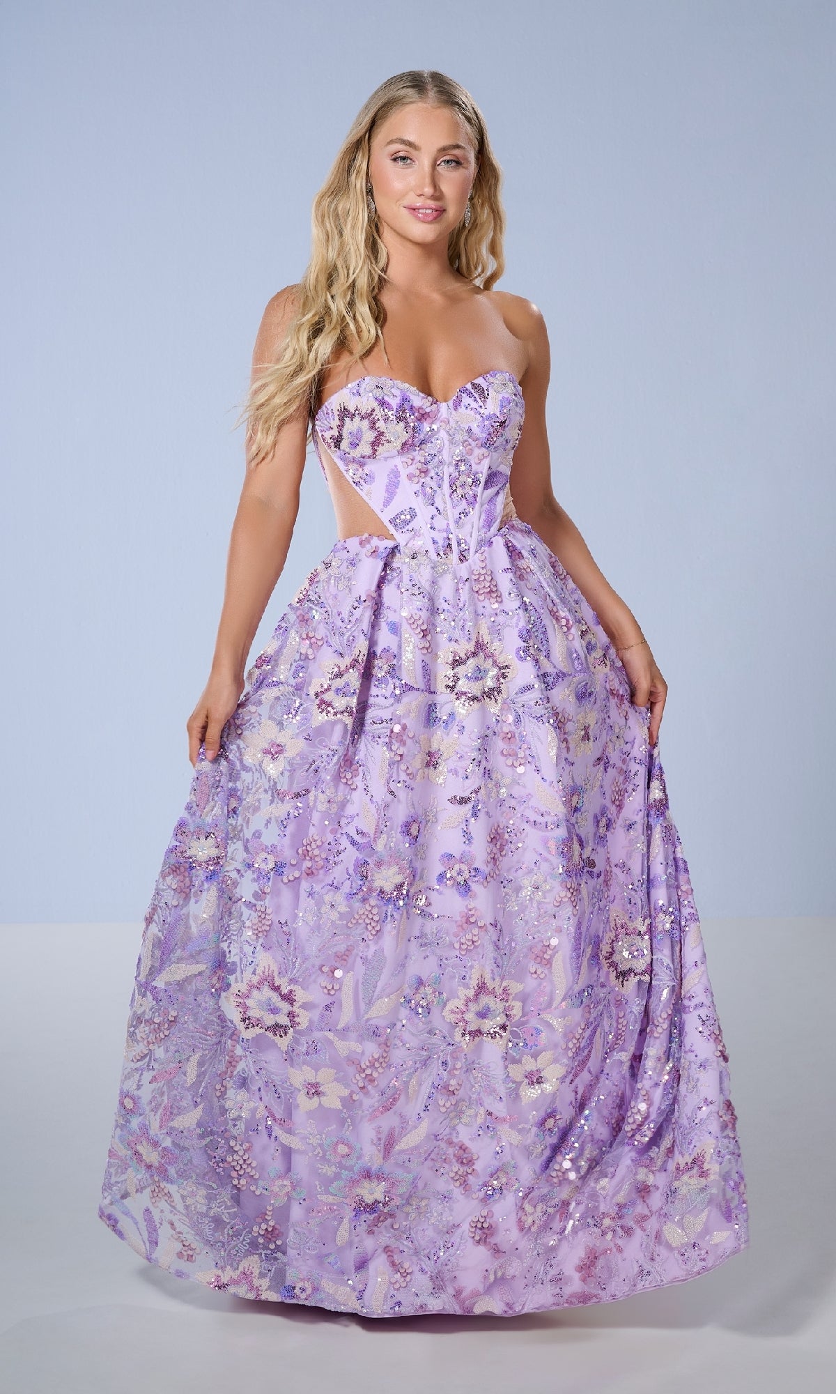 A woman stands against a plain light background wearing the Tiffany 16238 long prom dress—a strapless gown with a sweetheart neckline, cut-out sides, full skirt, and lavender floral patterns.